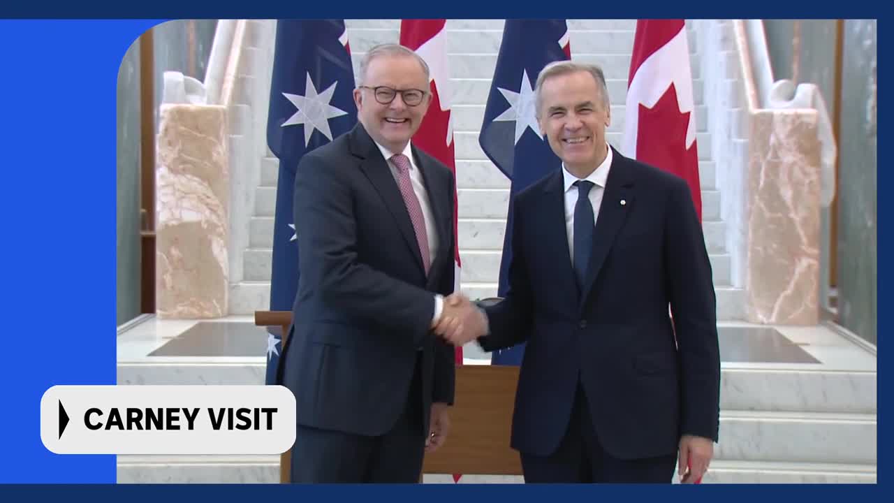 Two men in dark suits shake hands, their smiles wide. Behind them, Australian and Canadian flags stand tall.
