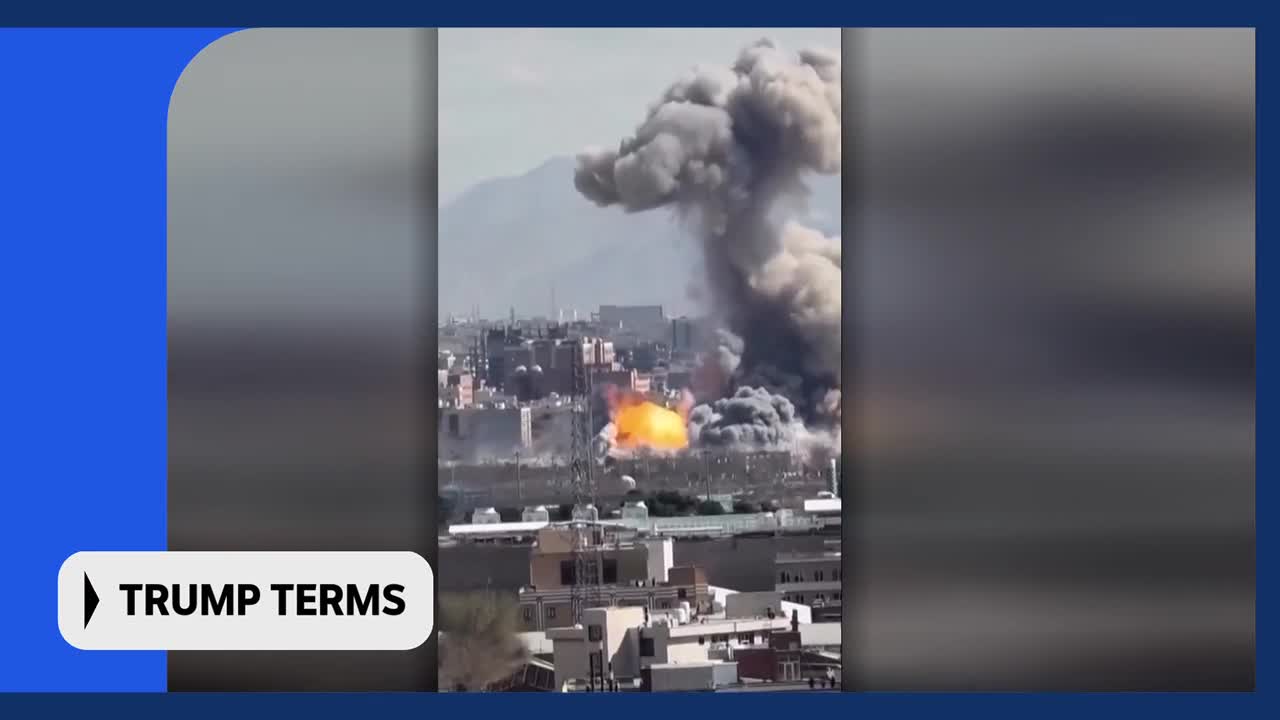 Thick grey smoke billows upwards from a city, obscuring buildings and mountains behind it. A bright orange explosion flares near the base of the smoke plume.
