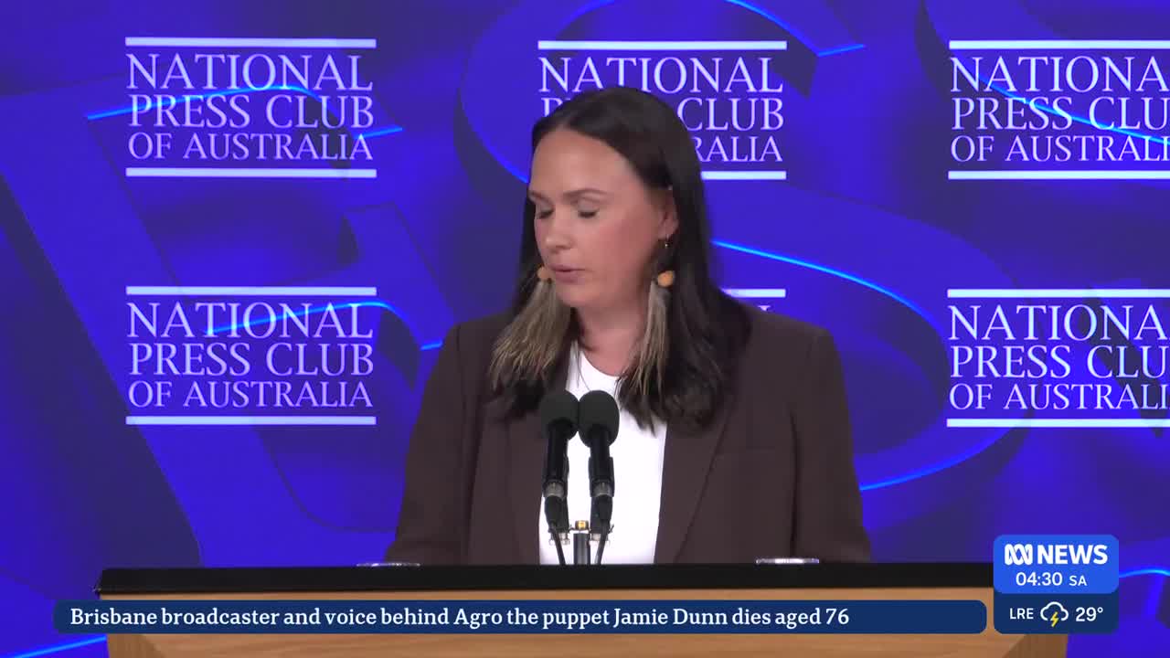 A woman stands at a podium, speaking into two microphones. The backdrop behind her is a deep blue with repeated logos for the National Press Club of Australia. A ticker at the bottom of the screen shows ABC News and a local weather report for South Australia.
