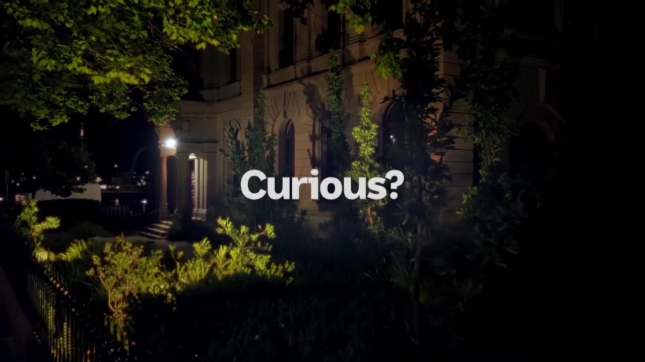 The grand facade of a building looms in the darkness, illuminated by strategically placed lights. A single word, "Curious?", hangs in the air, prompting a sense of intrigue.