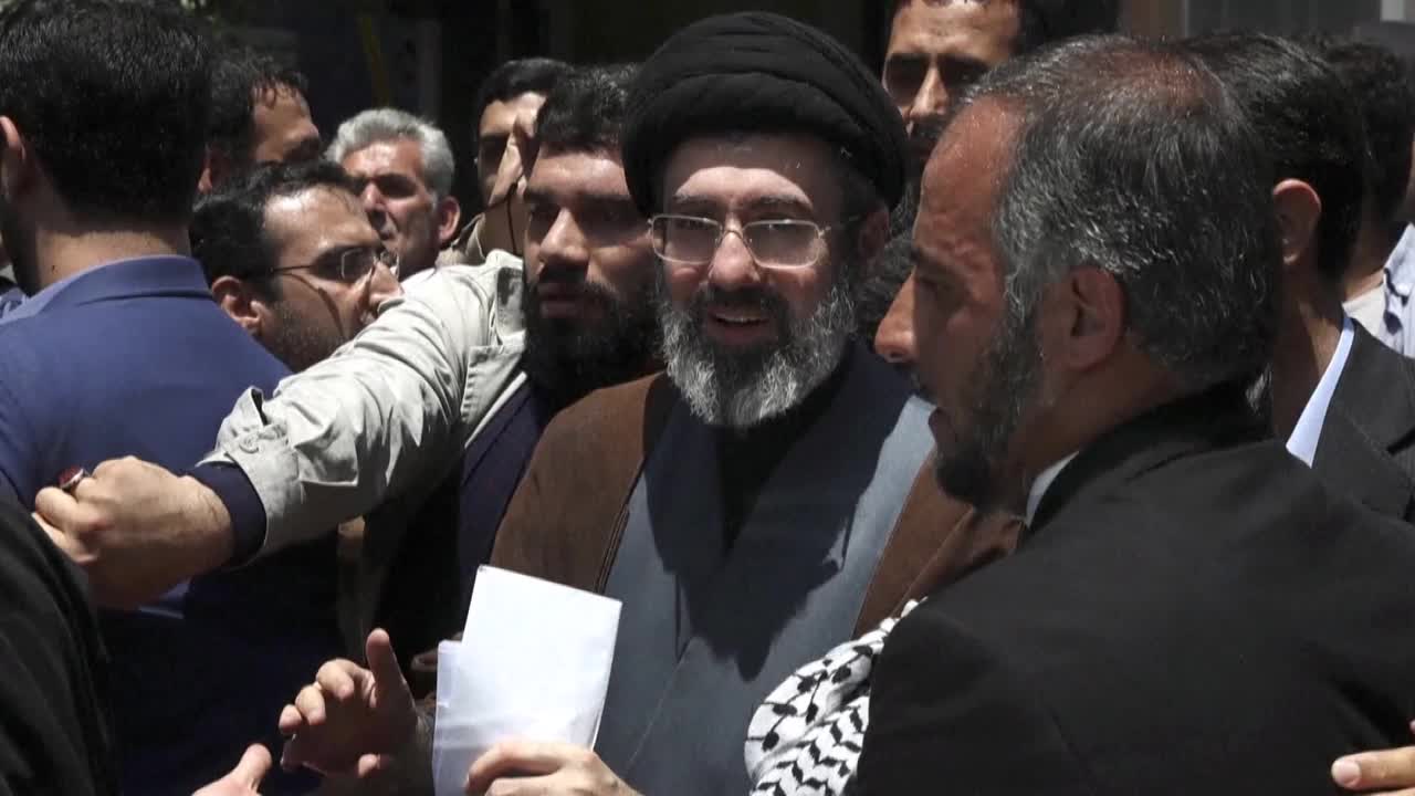 A man in a black turban and glasses smiles, holding white papers as a crowd presses in. Another man in a dark suit turns his head, his beard framing his profile.