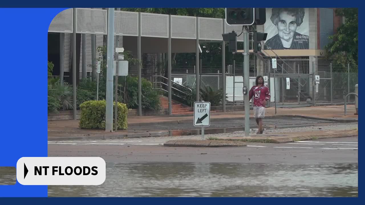 A person walks along a flooded footpath. The water is deep enough to cover the road in front of them.