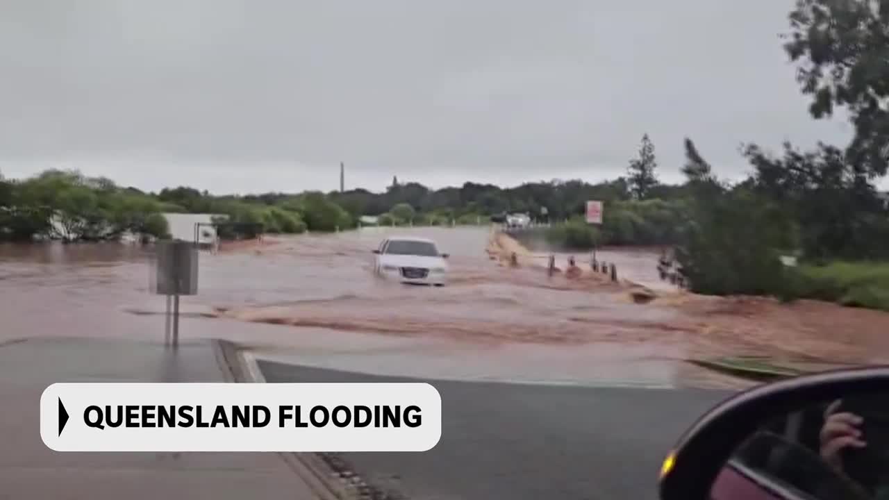 A white sedan is driving through a flooded road, its tires submerged in the brown water. The ABC Australia graphic at the bottom confirms this is Queensland flooding.