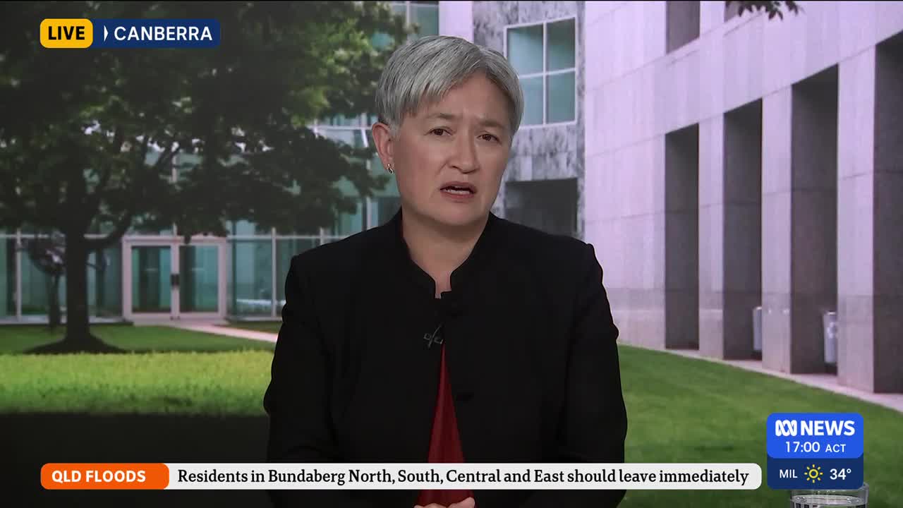 A woman in a black jacket speaks directly to the camera, her expression serious. Behind her, a modern building and manicured lawn suggest a government precinct in Canberra.