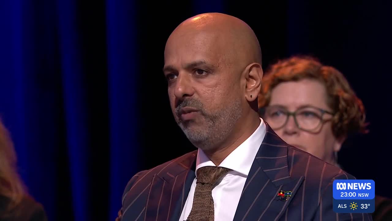 A man in a pinstripe suit speaks, his gaze directed slightly off-camera. Behind him, a woman with curly hair and glasses watches intently.
