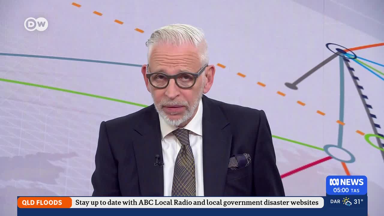 A man in a dark suit and patterned tie speaks directly to the camera. A graphic overlay at the bottom of the screen indicates "QLD FLOODS" and advises to stay updated with ABC Local Radio and local government disaster websites.