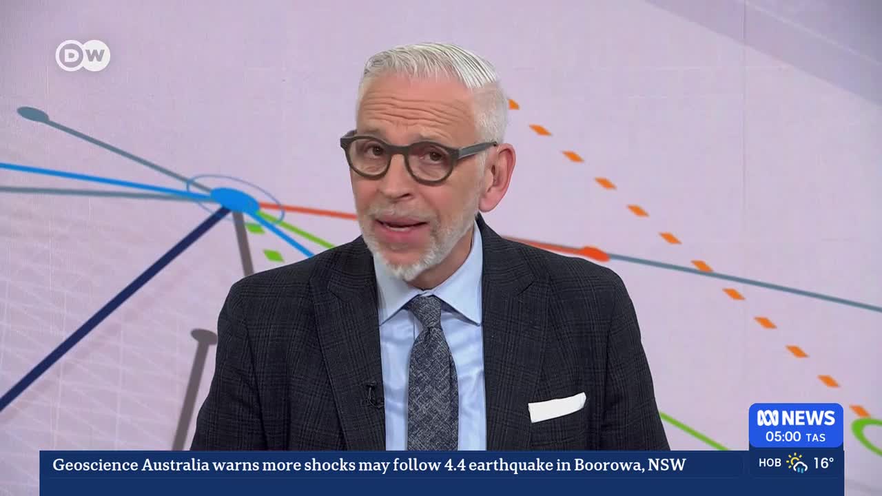 A man in a suit and tie speaks directly to the camera. A graphic behind him shows interconnected lines and dashed orange shapes. A news ticker at the bottom reports on an earthquake in Boorowa, NSW.