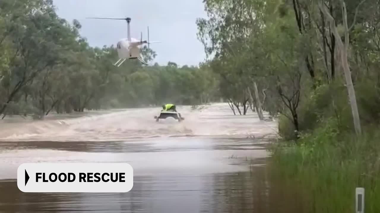 A small white helicopter hovers above a flooded road where a vehicle with a person in a bright yellow vest is navigating the churning water. This is a flood rescue operation happening somewhere in Australia, likely being reported by ABC Australia.
