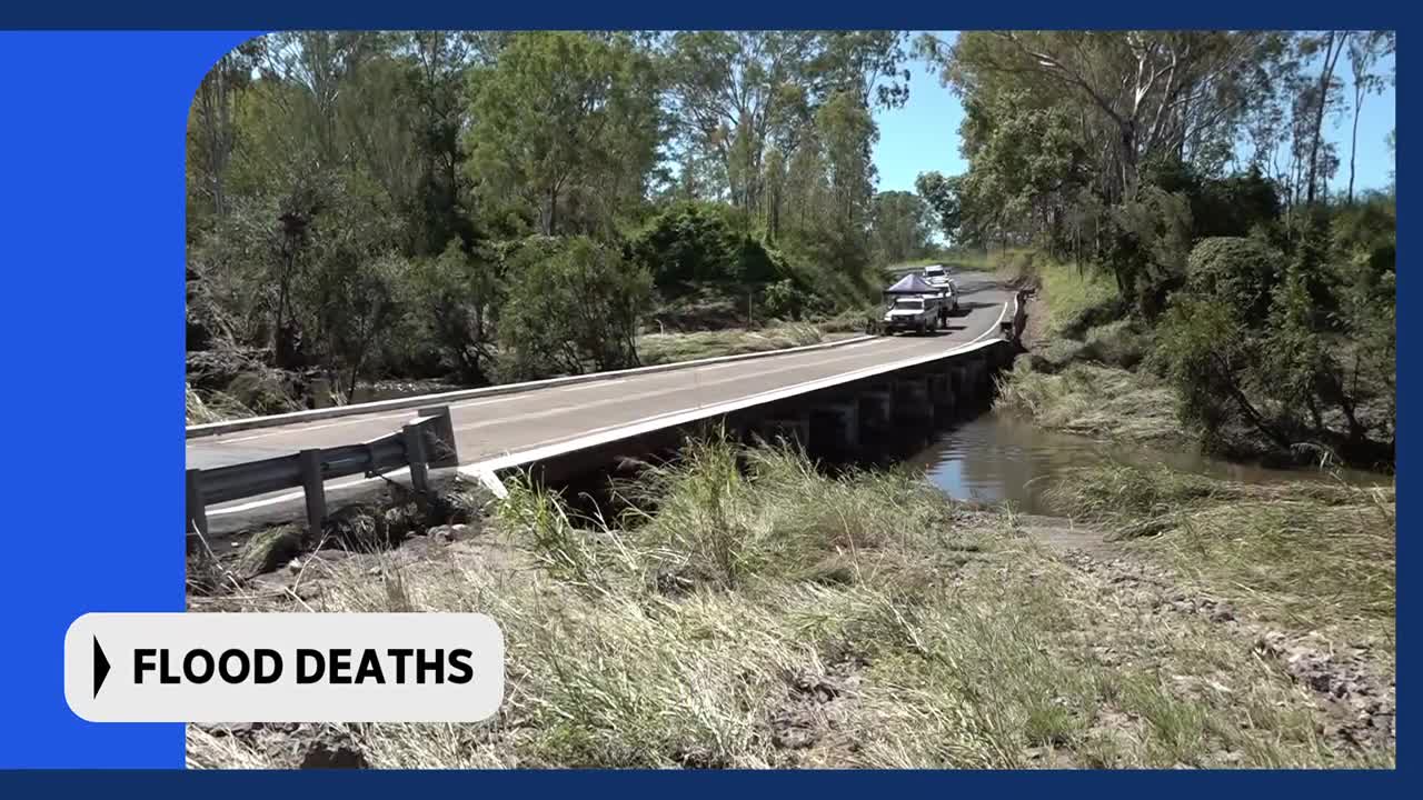 A white ute, carrying what looks like a camper trailer, drives across a concrete bridge. The water below is high, lapping at the banks and carrying debris.