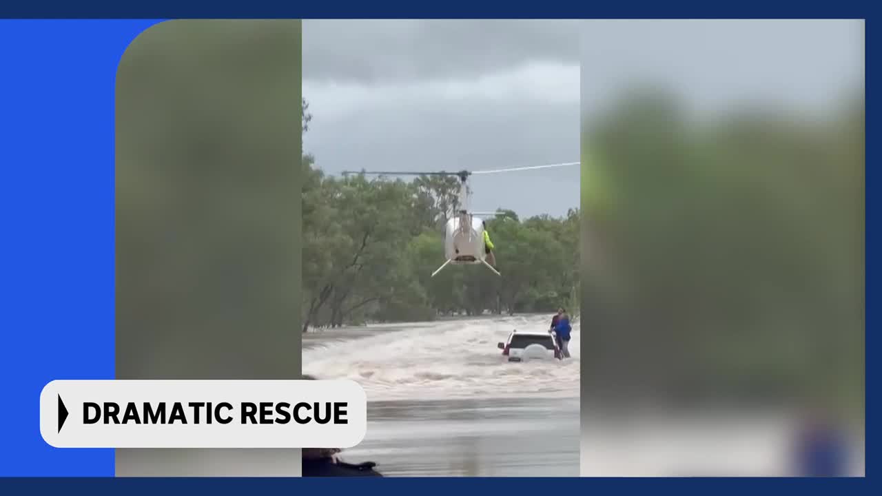 A helicopter hovers over a flooded road in rural Australia, its rotors churning the air. Two people stand near a submerged ute, waiting for assistance.