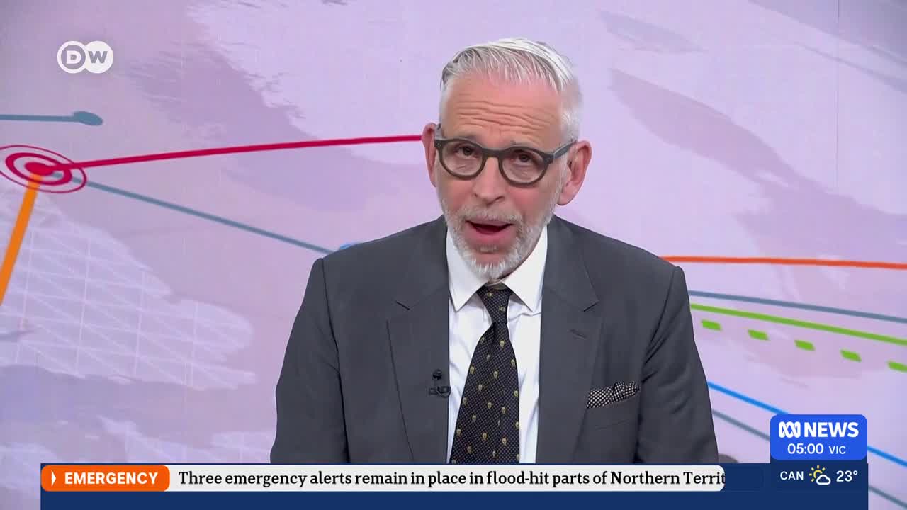 A man in a suit and tie speaks directly to the camera. Behind him, a graphic displays interconnected lines and circles, suggesting a map or network. A ticker at the bottom of the screen reports "EMERGENCY" and "Three emergency alerts remain in place in flood-hit parts of Northern Territory."