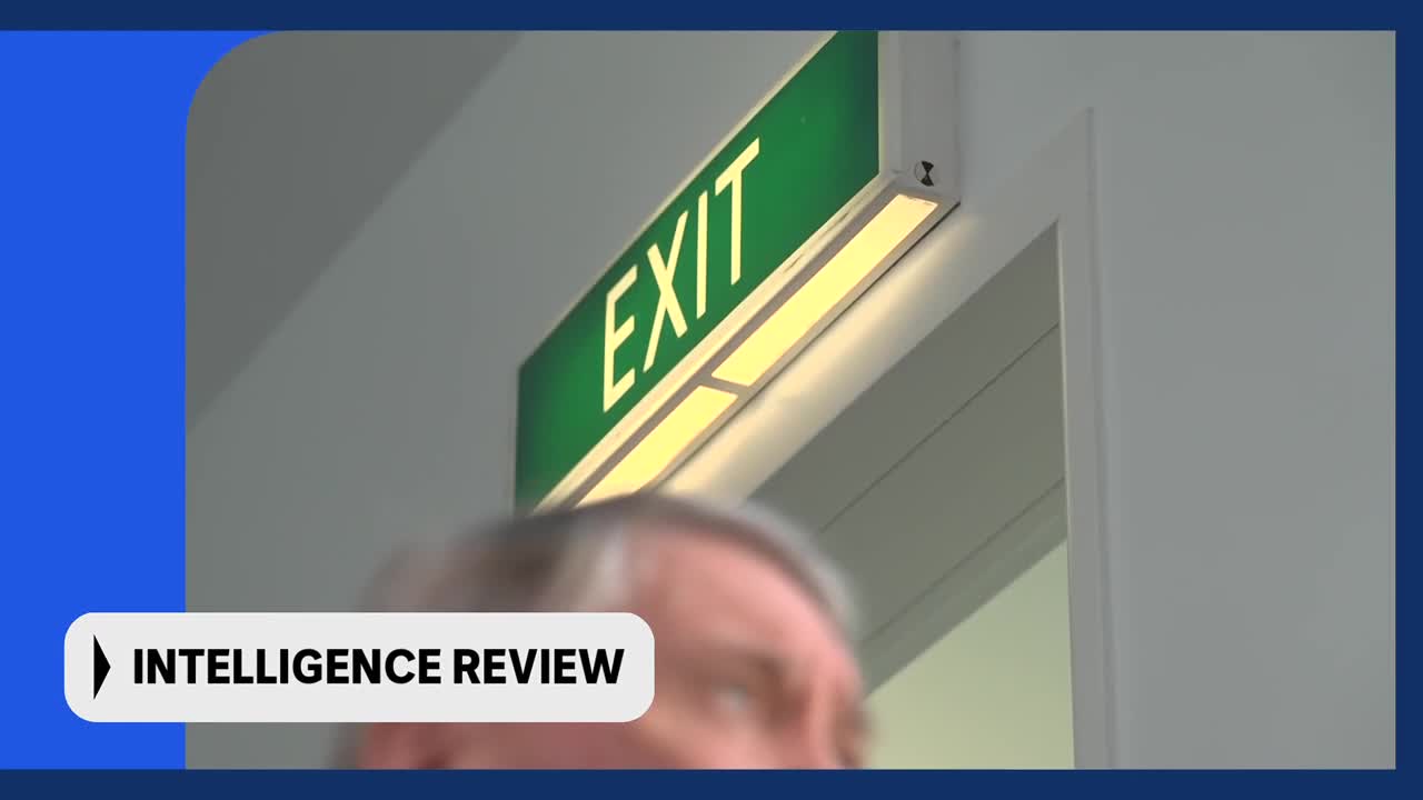 A man's head, blurred with motion, passes under a bright green "EXIT" sign. This is part of an "INTELLIGENCE REVIEW" segment, likely from ABC Australia.