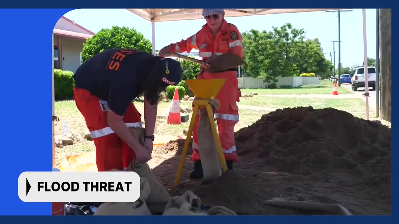 Two people in orange SES gear are filling sandbags from a large pile of dirt. The sandbags are being placed near a funnel on a tripod.