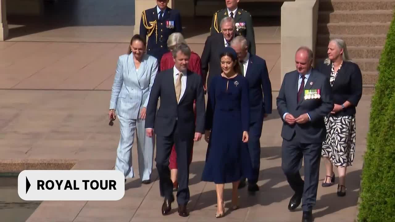 A group of people, including a man in a dark suit and a woman in a navy dress, walk together on a paved path. A banner in the corner reads "ROYAL TOUR." A group of people, including a man in a dark suit and a woman in a navy dress, walk together on a paved path. A banner in the corner reads "ROYAL TOUR."