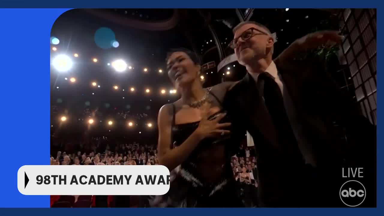 A woman in a black dress and a man in a suit are on stage. The audience is seated in the background, illuminated by stage lights. A woman in a black dress and a man in a suit are on stage. The audience is seated in the background, illuminated by stage lights.