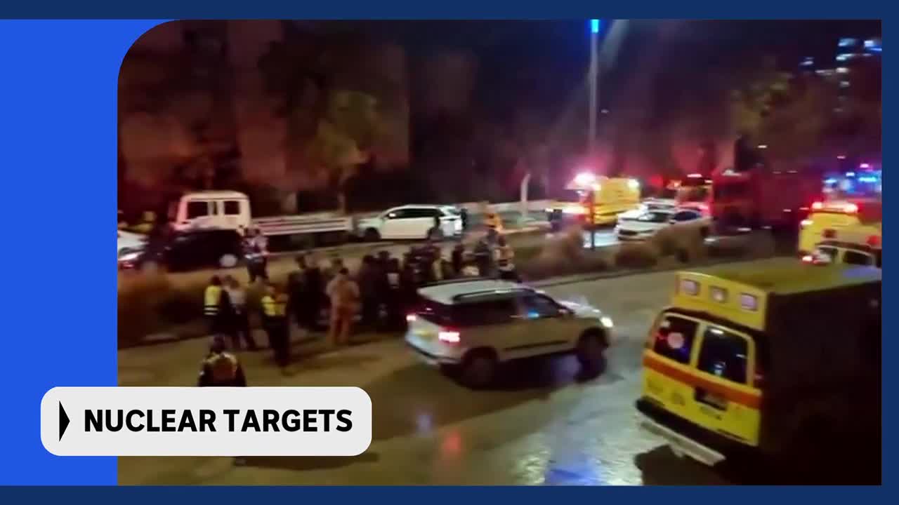 Emergency vehicles with flashing lights fill the street as a crowd gathers. A white SUV turns onto the road, its headlights cutting through the night.