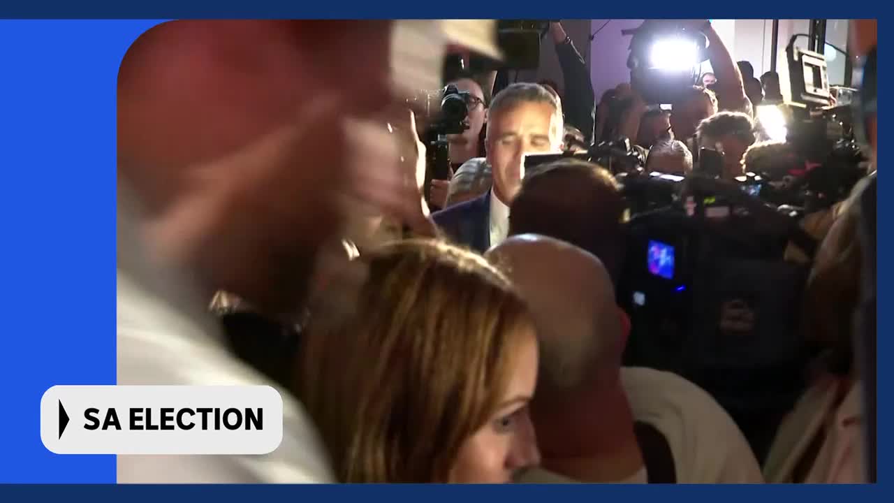 A man in a suit is being pushed through a crowd of people holding cameras and microphones. The lights are bright, and the energy feels electric, like after a big announcement on ABC Australia.