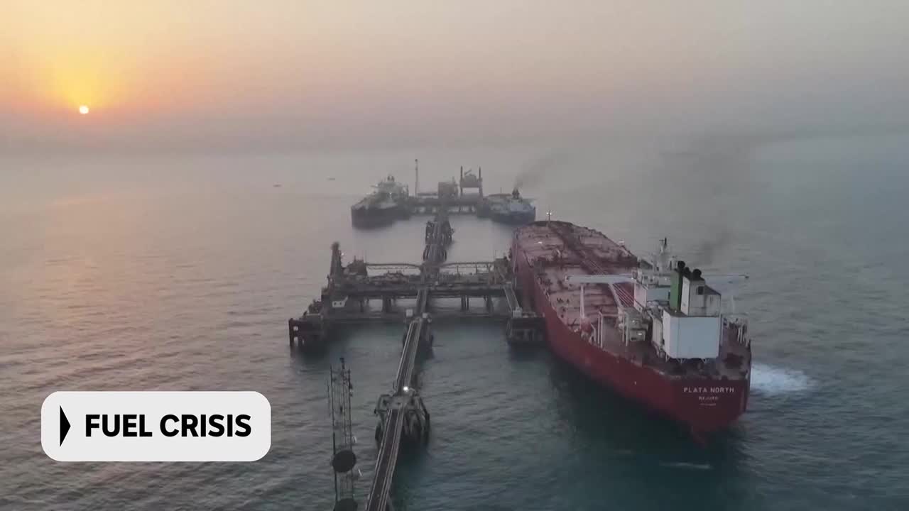 A large red tanker is docked at an offshore platform, its engines churning white foam into the calm water. Two other ships are moored further out, their stacks emitting faint plumes of smoke against the hazy sunrise.