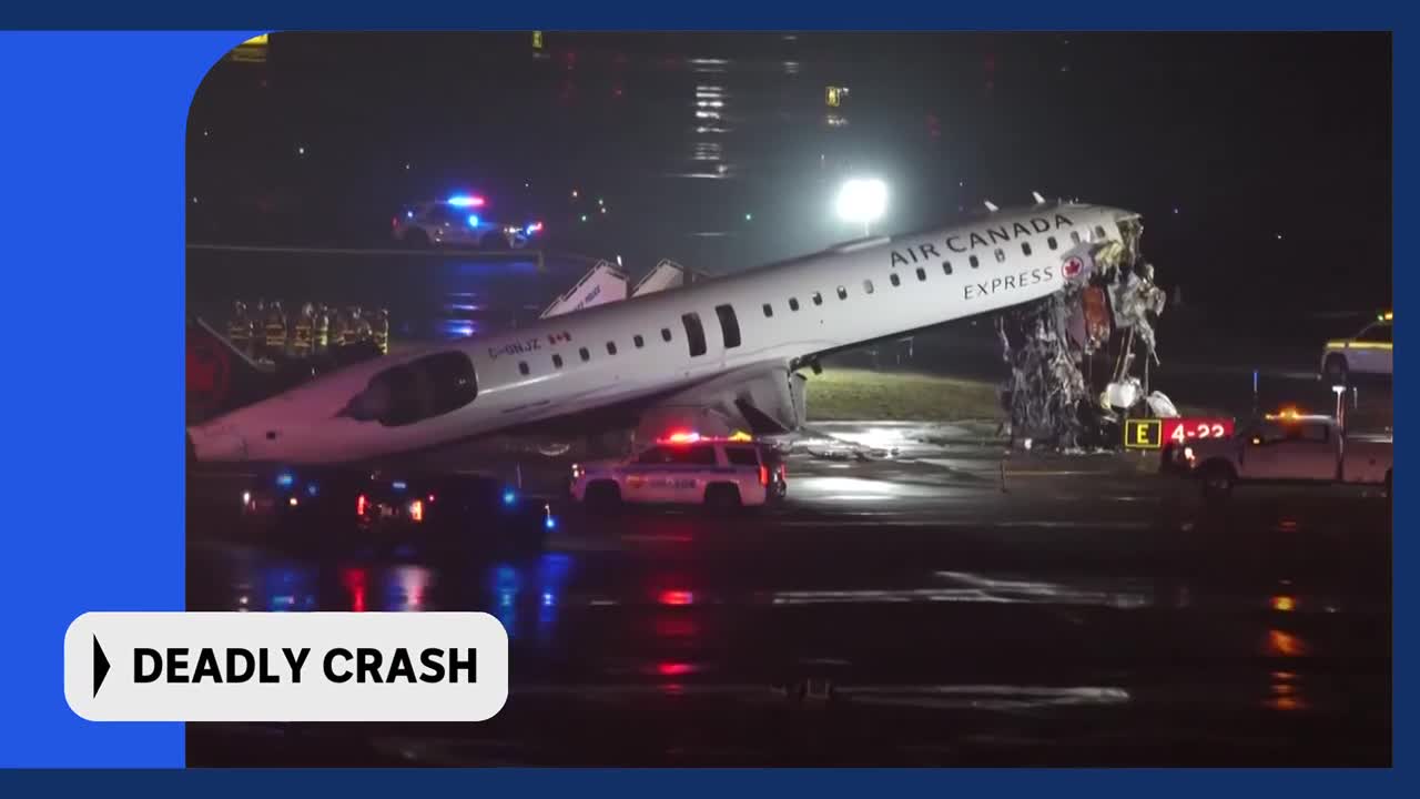 An Air Canada Express plane is tilted on its side on a wet tarmac, its tail section severely mangled. Emergency lights flash from police vehicles and what appears to be a fire truck as personnel gather near the wreckage.