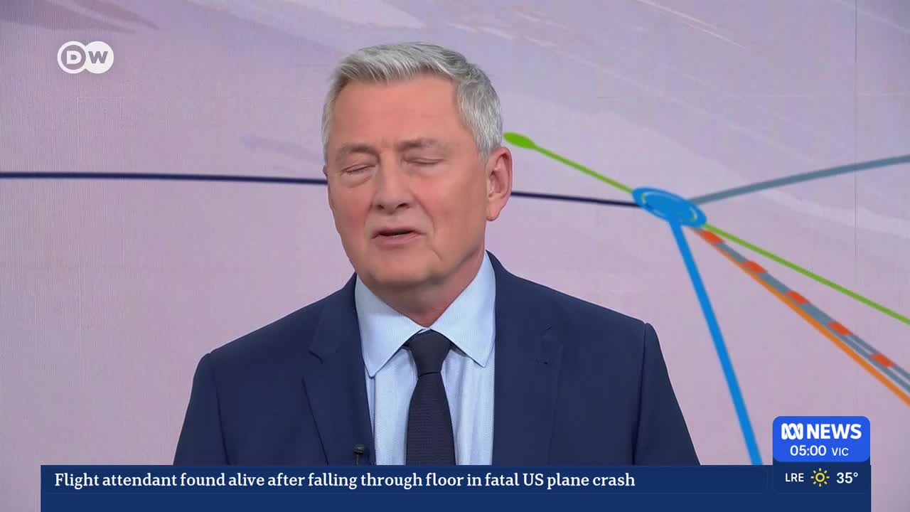 A man in a dark suit and tie speaks directly to the camera. Behind him, a graphic displays a network of coloured lines.