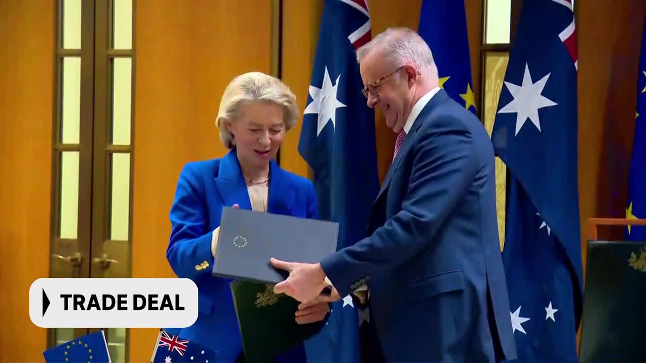 The Australian Prime Minister and the European Commission President are exchanging documents. Australian flags stand behind them as they finalize a trade deal.
