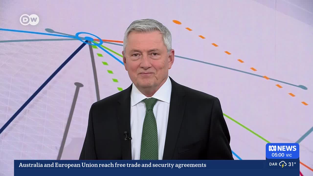 A man in a suit stands before a screen displaying a network of colored lines. The ABC Australia news ticker at the bottom reports on agreements between Australia and the European Union.