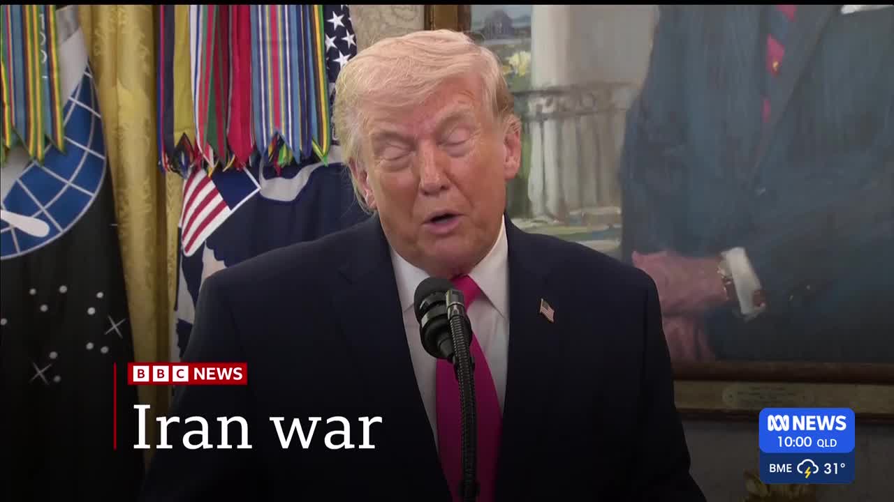 The man in the suit speaks into a microphone, his eyes closed. Behind him, a row of colourful medals hangs above flags. A news ticker from ABC Australia shows the time and temperature.