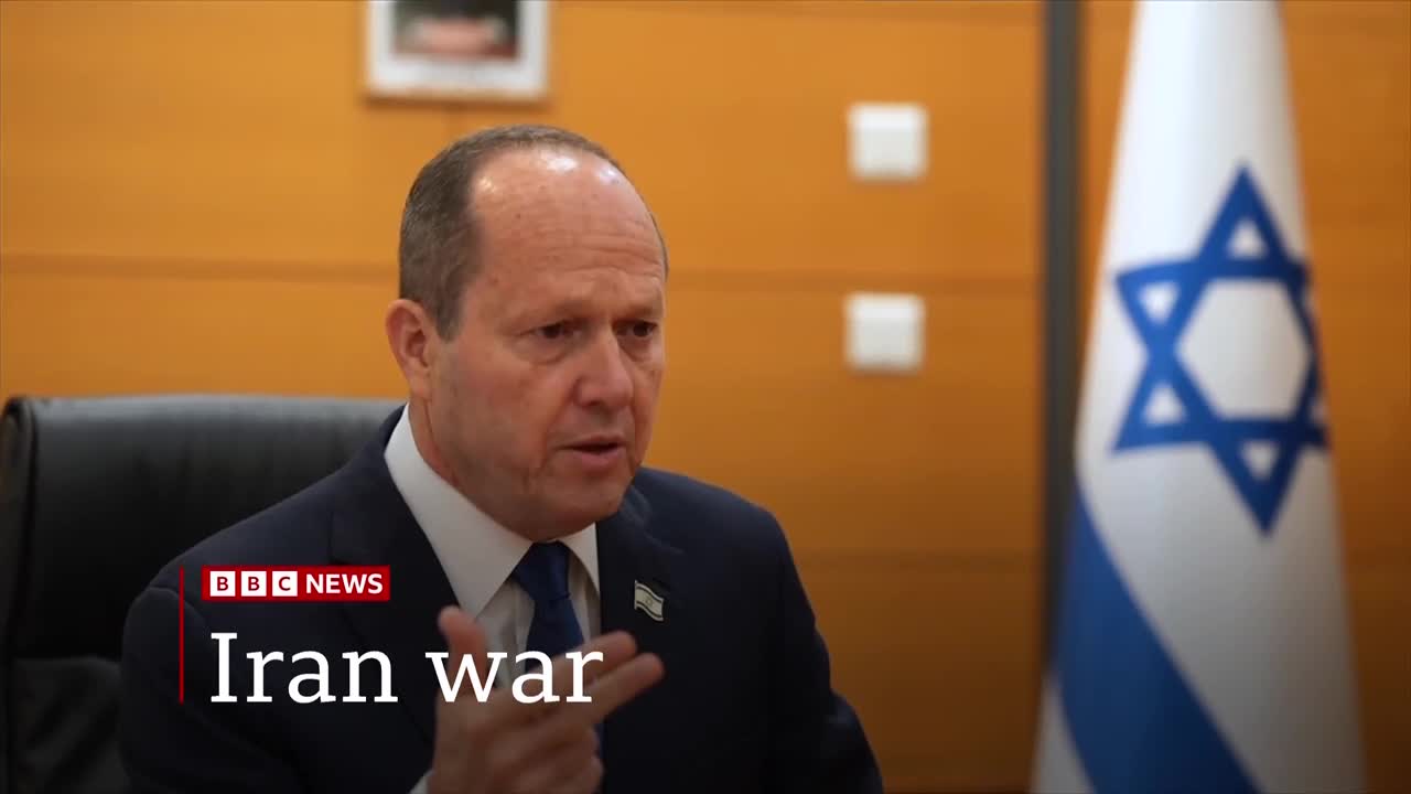 A man in a suit speaks, gesturing with his hands. Behind him, the Israeli flag stands tall.