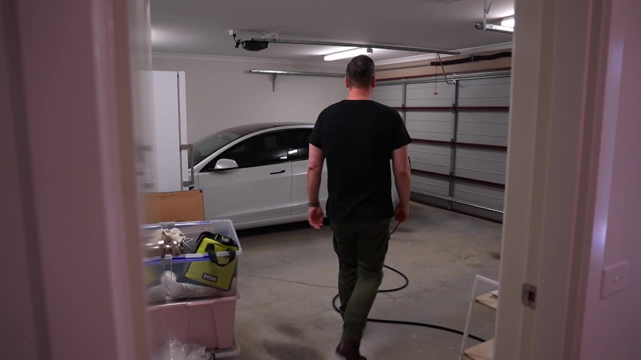 A man walks into a garage towards a white electric car. He's carrying a black cable, likely for charging.
