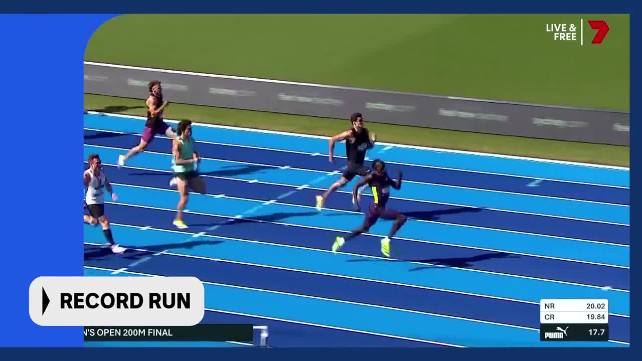 The runners are nearing the finish line in the men's 200m final. One athlete in a dark purple uniform pulls ahead, his arms pumping as he sprints towards the tape.