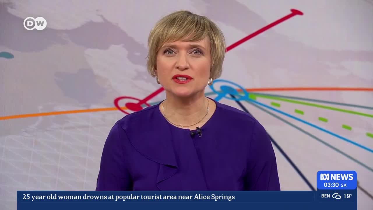 A woman in a purple top speaks directly to the camera, her blonde hair neatly styled. Behind her, a graphic displays a network of coloured lines radiating outwards. A news ticker at the bottom of the screen reports a drowning near Alice Springs.