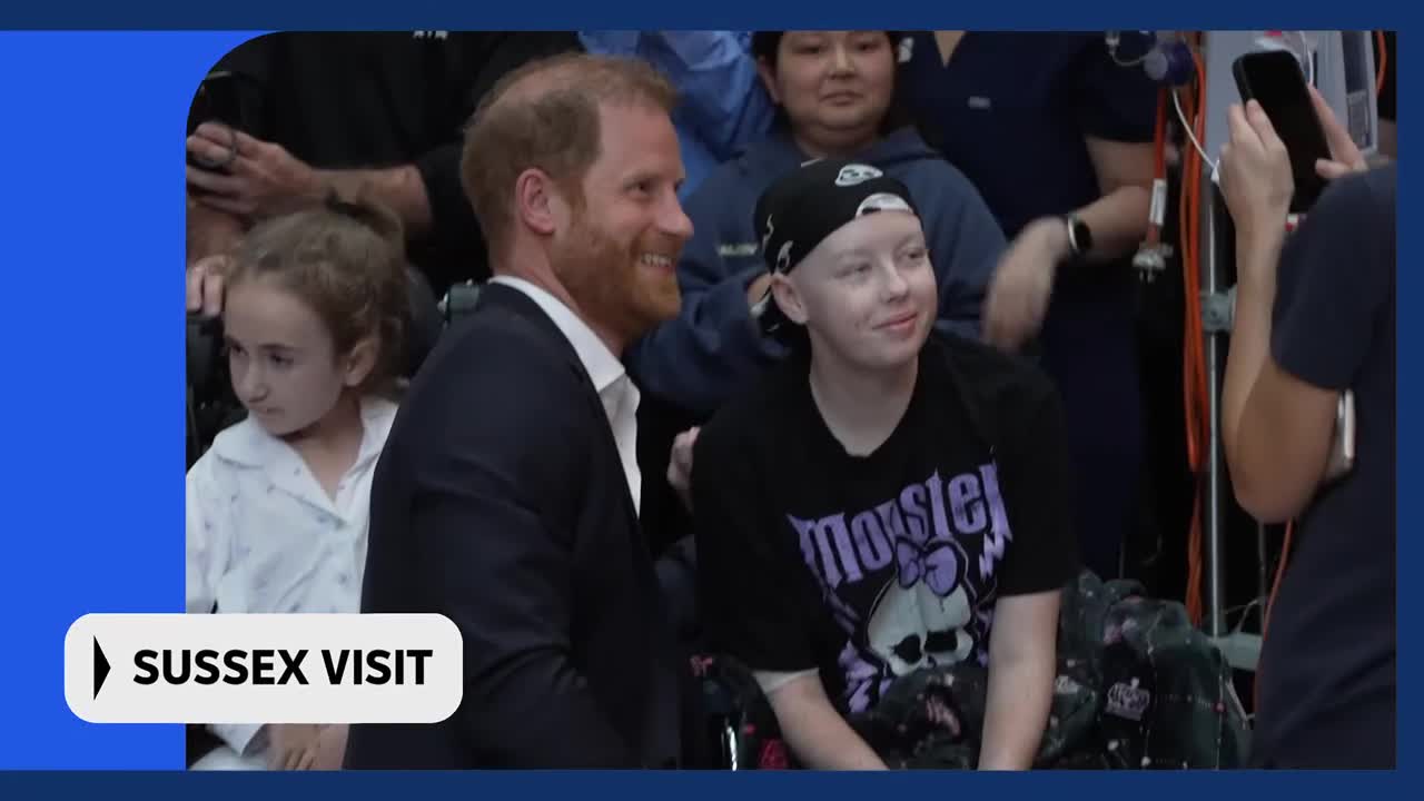 Prince Harry smiles as he speaks with a young girl wearing a bandana and a "Monster" t-shirt. A child in a white shirt sits beside them, looking on.