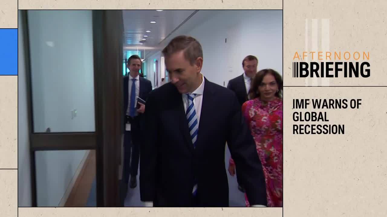 A man in a dark suit and a blue striped tie walks down a hallway, looking down. Behind him, two other men in suits and a woman in a bright floral dress follow.