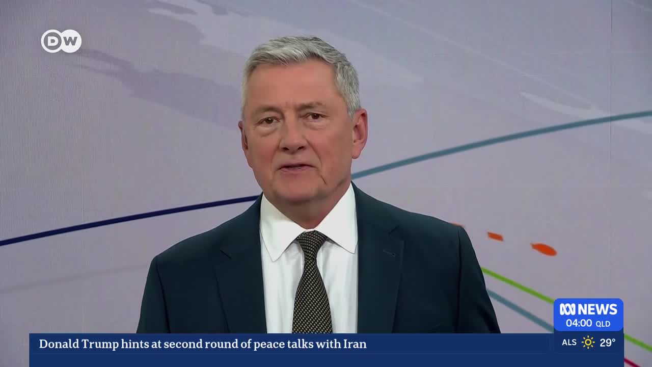 A man in a dark suit and tie stands before a screen displaying abstract lines and shapes. At the bottom, ABC News text indicates the time and temperature in Queensland.