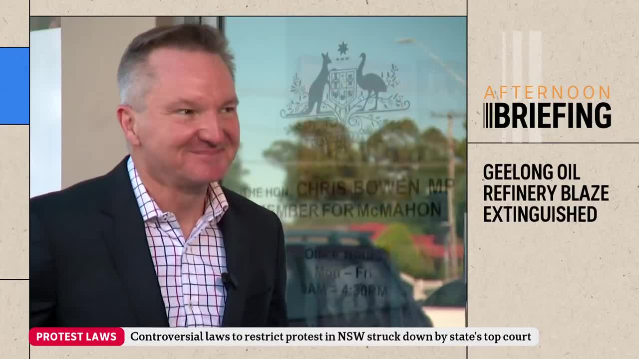 A man in a suit jacket smiles at the camera. Behind him, a glass door reflects the Australian coat of arms and text that reads "The Hon. Chris Bowen MP Member for McMahon."