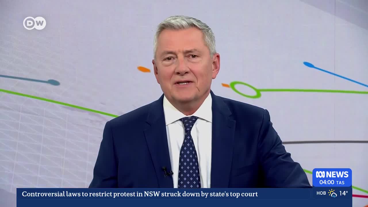 A man in a suit speaks directly to the camera. Behind him, colorful lines curve across a white background, and a ticker displays news from ABC Australia.