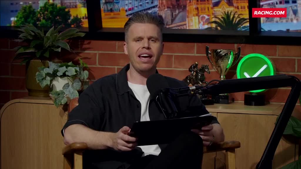 A man in a black shirt is speaking, holding a tablet. He's sitting in a wooden chair, in a studio with a Racing.com logo visible.
