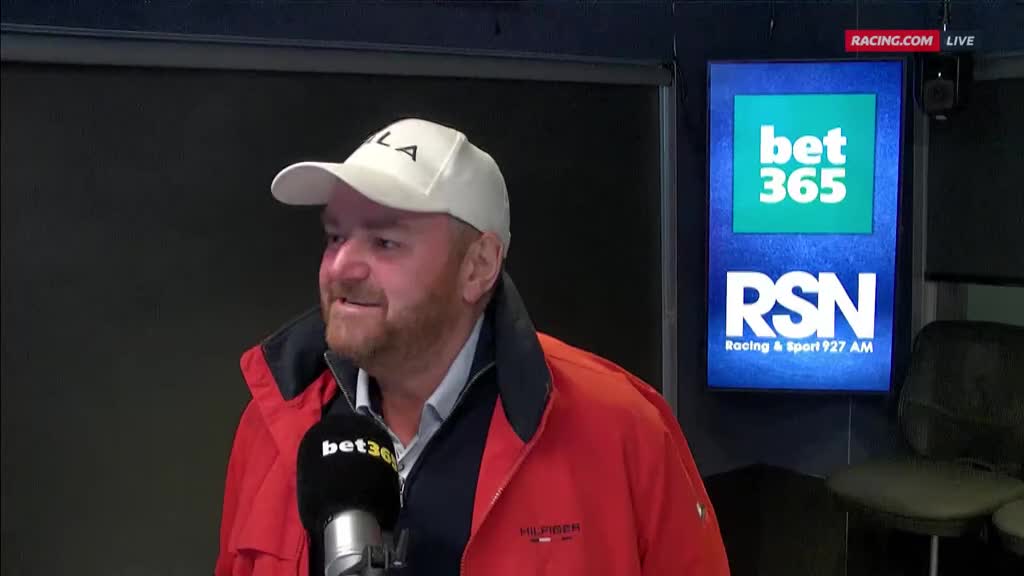 The man in the white cap and red jacket is speaking into a microphone, the bet365 logo visible on it. Behind him, a screen displays the Racing & Sport 927 AM logo, and the Racing.com website address.
