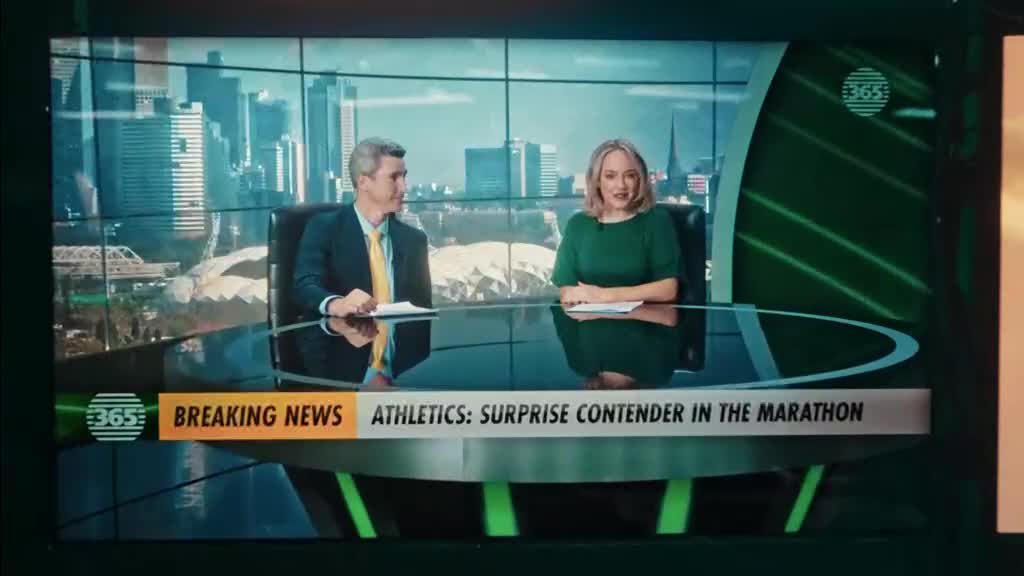 Two news presenters sit behind a curved desk, the backdrop a cityscape. The screen flashes "Breaking News" with a headline about a surprise contender in the marathon.
