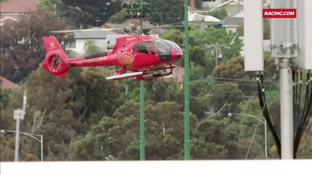 A bright red helicopter, emblazoned with a logo, hovers above the trees. The Racing.com broadcast is underway, and the chopper is likely capturing the action from above.
