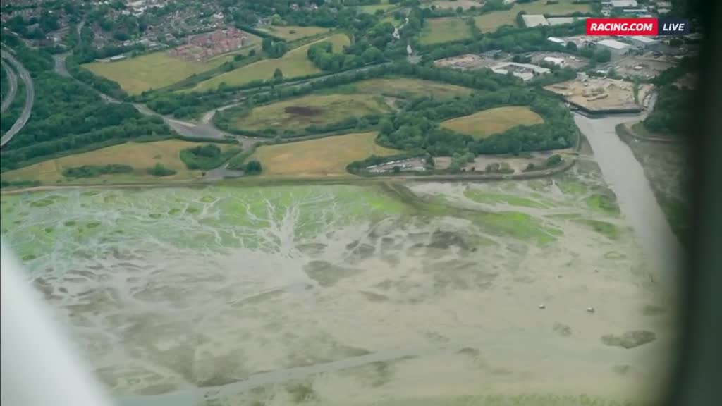 From this high vantage point, I see a network of pale, watery channels snaking through a green and muddy expanse. The land rises to fields and trees, with a town visible in the distance, all under the Racing.com banner.

