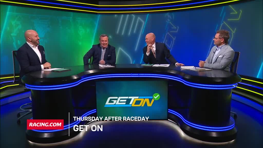 Four men in suits are seated at a curved desk, the Racing.com logo visible below. They're on the "Get On" program, and they're all smiling and chatting.
