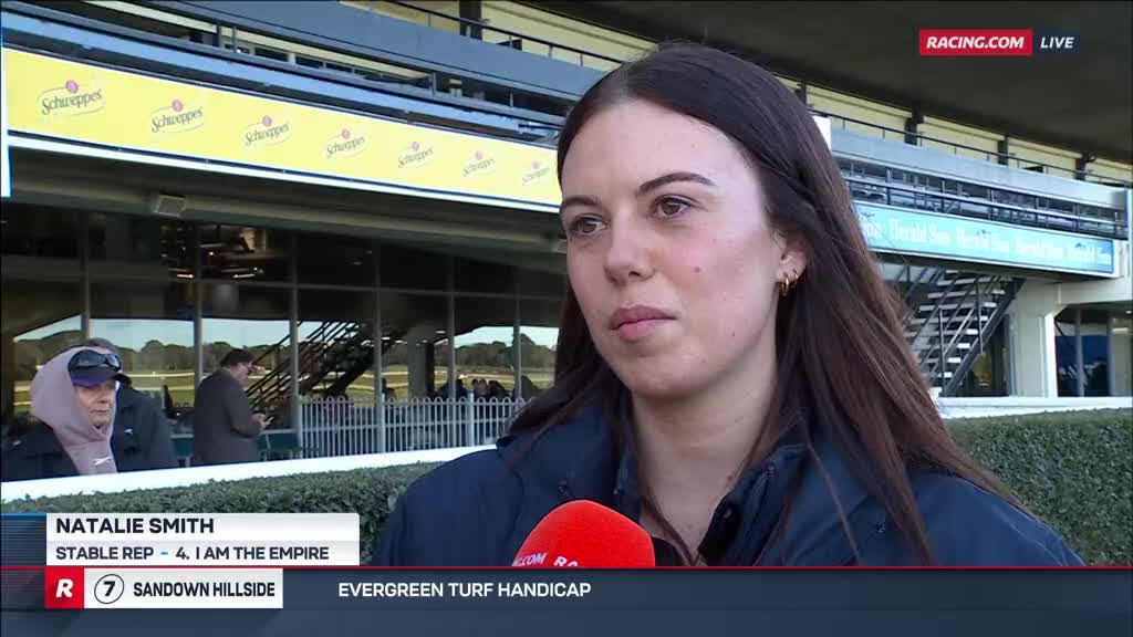 Natalie Smith, a stable rep, is being interviewed at Sandown, with the Racing.com logo visible. She's standing trackside, a red microphone held near her, with the stadium and its signage in the background.
