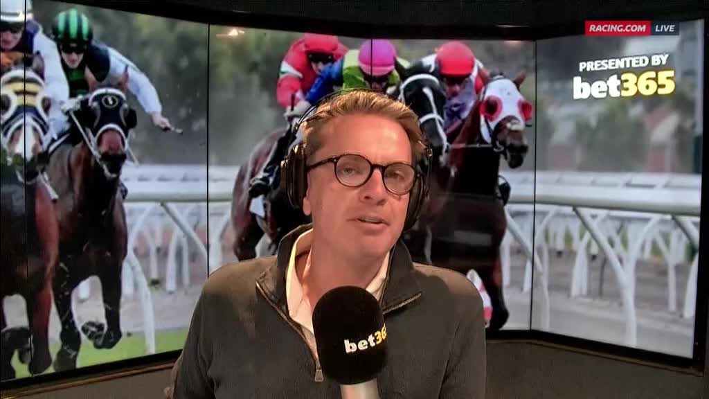 The commentator, wearing glasses and headphones, is speaking into a microphone branded with "bet365." Behind him on Racing.com, horses with jockeys in colorful silks are racing.

