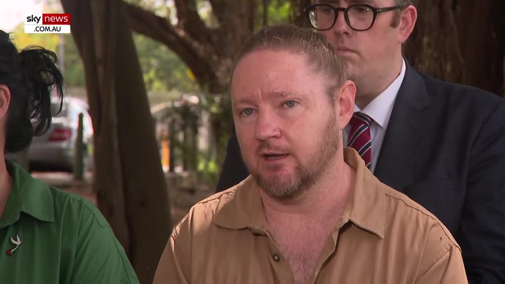 A man in a tan shirt speaks, his hair pulled back in a bun. Behind him, a man in a dark suit and striped tie stands attentively.