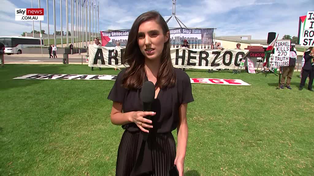 Protesters hold banners in front of Parliament House in Canberra. A Sky News Extra reporter stands with a microphone, speaking to the camera.