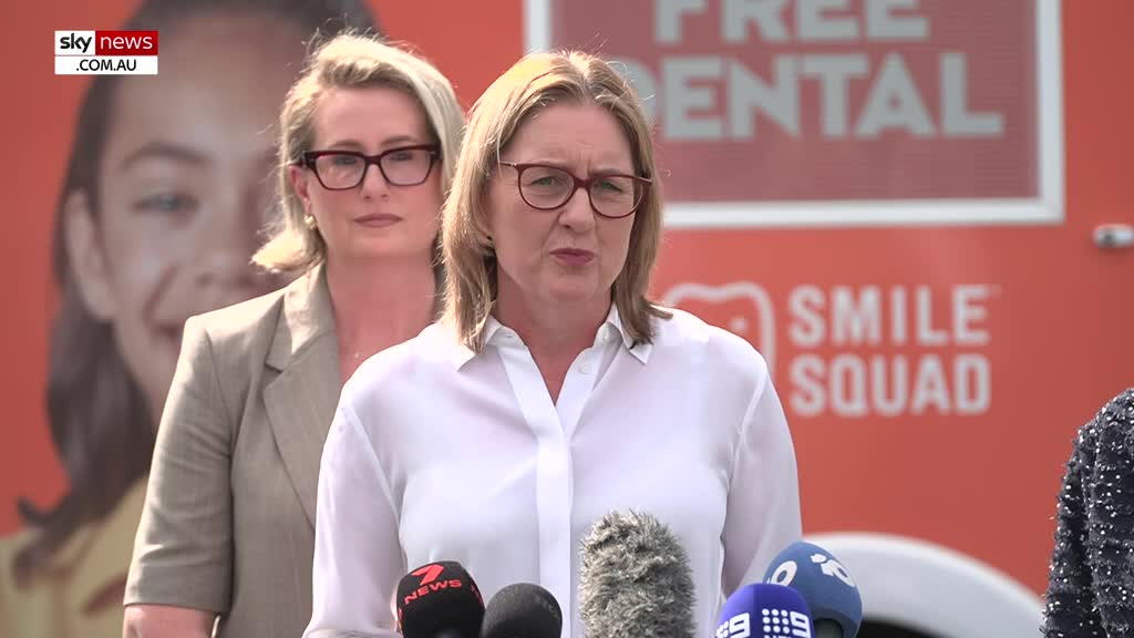 A woman in a white shirt speaks into microphones, with a large graphic behind her that reads "SMILE SQUAD." Another woman stands behind her, looking towards the side. A woman in a white shirt speaks into microphones, with a large graphic behind her that reads "SMILE SQUAD." Another woman stands behind her, looking towards the side.