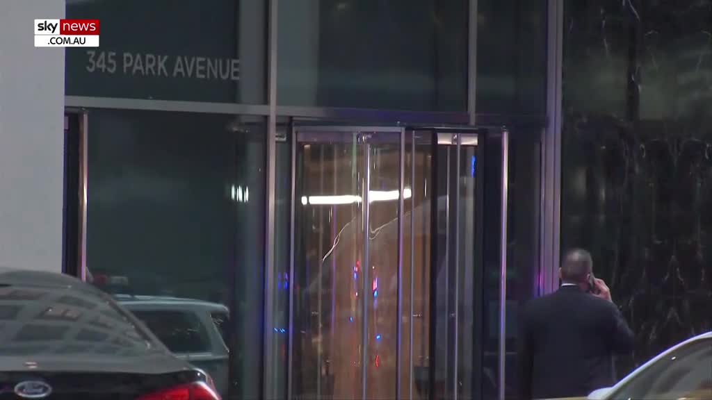 The revolving doors of 345 Park Avenue are turning slowly, reflecting the streetlights. A man in a suit is on the phone, standing near the entrance.
