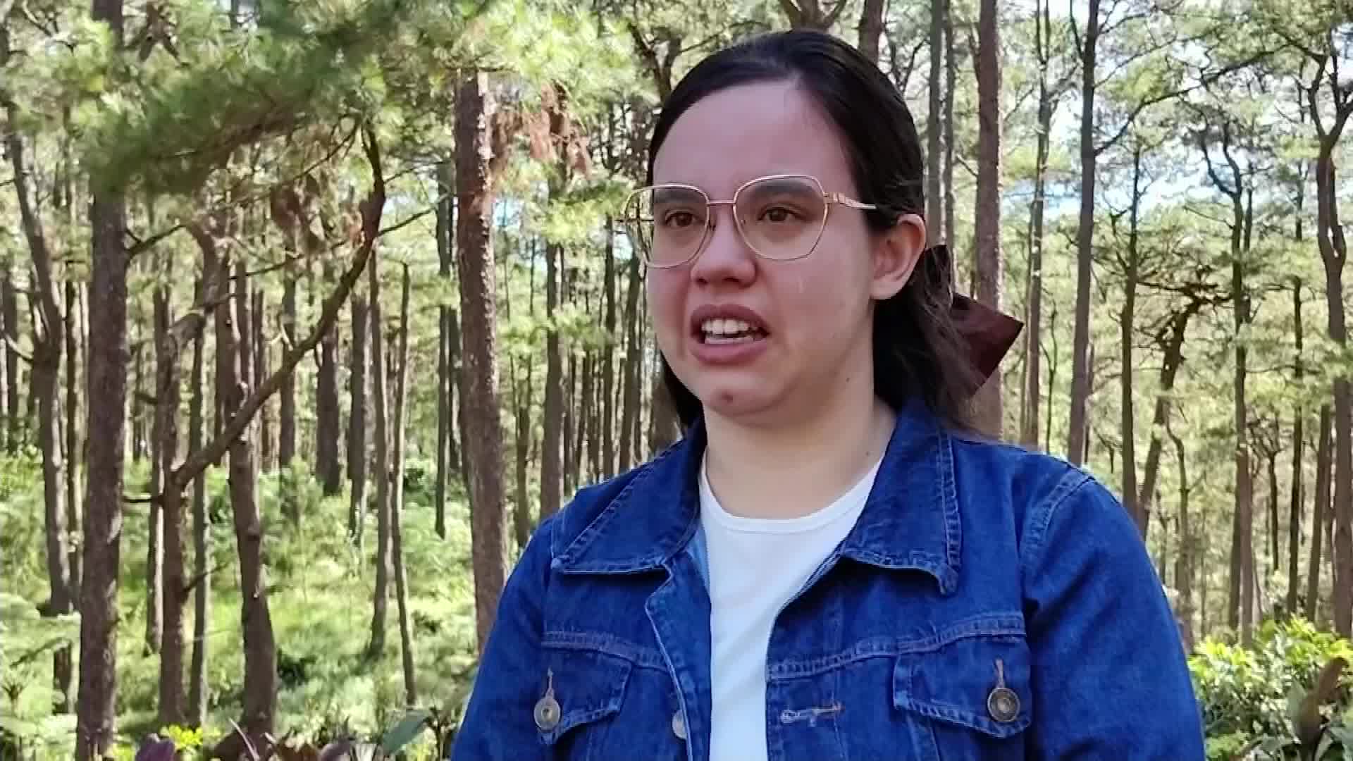 A woman in a denim jacket and glasses speaks, her mouth open as if mid-sentence. Behind her, tall trees create a forest backdrop.
