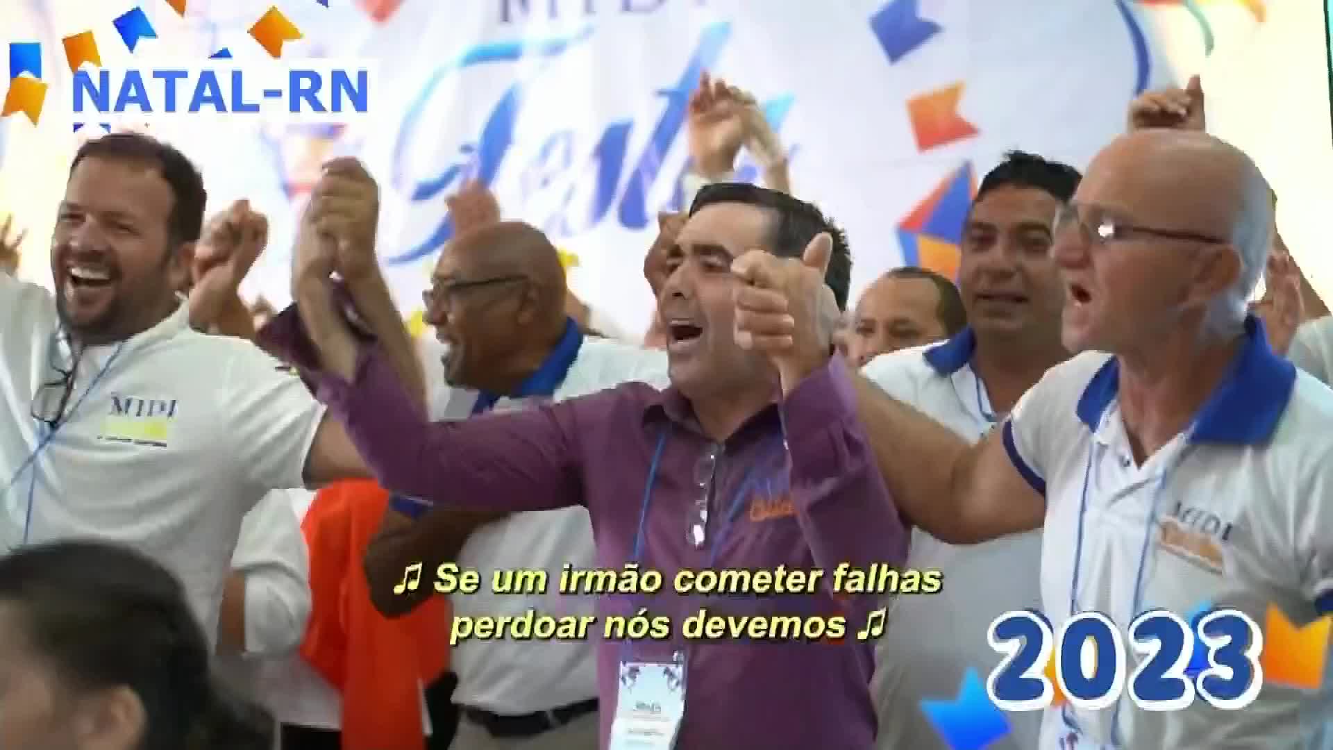 A group of men, all in white shirts with blue trim, are singing and raising their arms. One man in a purple shirt gives a thumbs-up as they celebrate.
